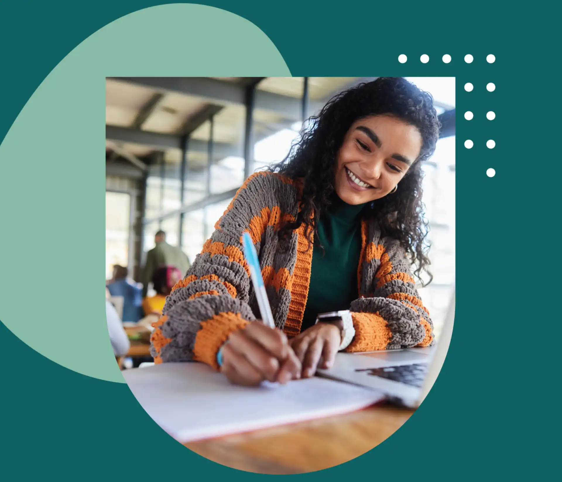 A young woman smiling while taking notes on paper with her laptop open.